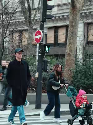 The image shows a street scene in an urban environment on what appears to be an overcast day. A man wearing a black coat and a baseball cap is walking on a crosswalk, with a woman pushing a baby stroller and a baby in the stroller wearing a pink hat. Behind them, in the background, are other people, buildings and a traffic light.