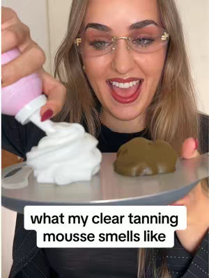 A woman with blonde hair, gold rimmed glasses, and red lipstick is holding a silver tray with two piles of product - one white fluffy cloud, and one brown opaque glob on it. She has a pink aerosol bottle in her other hand, presumably dispensing the white product. Below the tray, the text reads "what my clear tanning mousse smells like". The woman is looking up and smiling broadly.