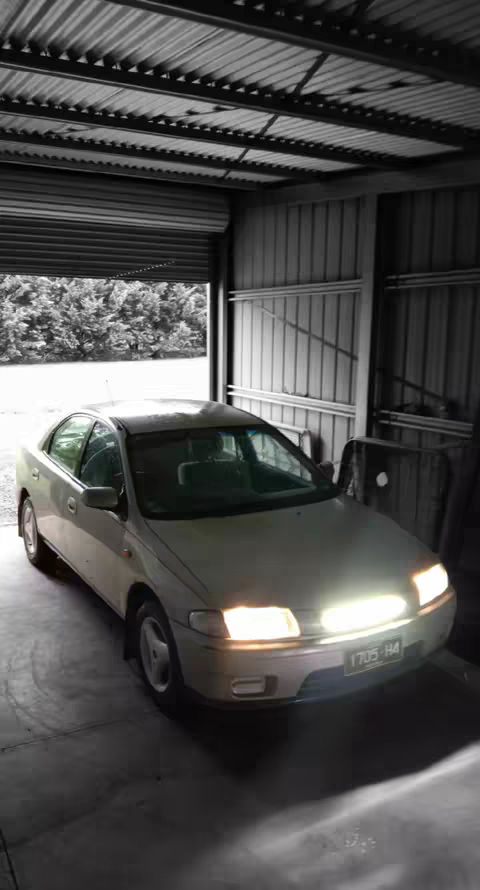 A monochrome image of a car parked inside a garage or a similar enclosed structure. The car is a light color, and its headlights are illuminated. The garage has a metal or corrugated roof and walls. The background includes a glimpse of trees.