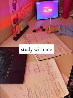 The image shows a desk setup for studying. There's a laptop on the left, a tablet in the center displaying a timer set to 11:00, papers covered in notes, and highlighters neatly arranged. The overall composition conveys a study environment with a focus on productivity. The background has pink and yellow lighting.