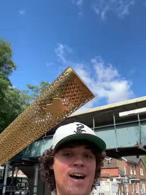 A selfie-style shot shows a young man in a baseball cap, smiling with an open mouth. He is holding the Olympic torch in place against a background of a blue sky, green trees and architectural details, with a clear, bright day. 
