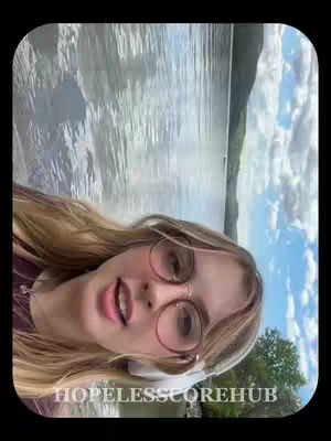 A young woman with long blonde hair poses for a selfie in front of a body of water with mountains visible in the background. She wears glasses and a burgundy colored shirt.