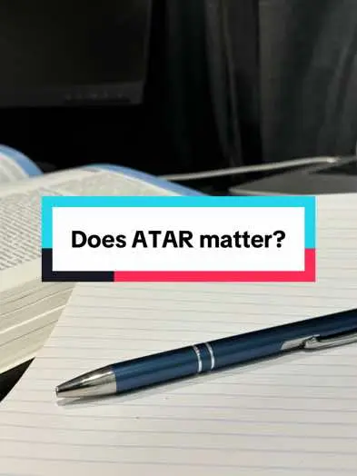 The image is a close-up shot of a pen on a lined paper. A blue pen is positioned diagonally, with the pen point in focus. The paper has lines, and on the left side, a book is partially visible with its pages open. A teal and red banner is placed in the center, the words "Does ATAR matter?" are in bold font. The background is blurred, suggesting depth. The color tones are simple, with the blue pen and banner colors contrasting against the white paper.