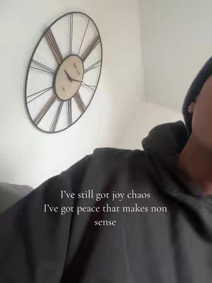 An image appears to be a selfie with background, the creator is shown with a dark hoodie, on a white wall there is a big clock.