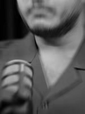 The cover image is a blurry, black and white shot from the chest up of a man holding a microphone. The man's face is out of focus, but a portion of his beard, jawline, and the microphone he's holding are discernible. The background is dark, and the overall effect is one of a close-up interview or podcast setting.