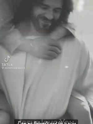 A grayscale image shows a man embraced from behind by another person, both set against a soft-focus background. The person in front, with visible facial features, appears to be smiling warmly. The composition emphasizes closeness and tenderness.