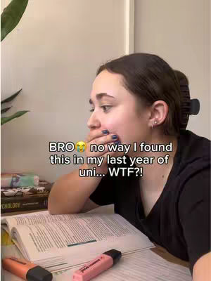 A woman is sitting at a table with a book open in front of her. She is looking down with a shocked expression, and her hand is covering her mouth. In the background, there is a shelf with some books.