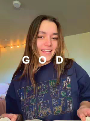 A young woman with long, light brown hair smirks at the camera, with the word "GOD" placed vertically over her face in a bold white font. She wears a dark blue t-shirt featuring a colorful design. The background has a light-colored ceiling and a wall with a few string lights.