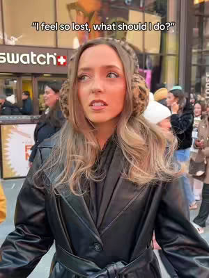 A woman with long, wavy blonde hair and leopard print ear muffs stands outside of a building. She wears a black leather coat and pauses to look slightly upwards with a concerned expression. In the background, other people walk by, and part of a sign is visible.