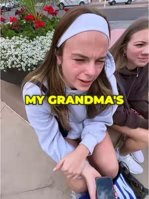 A young woman with long brown hair and a white headband is the central focus of the image. She is seated and leaning forward, her expression conveying distress or an emotional response, possibly crying. Beside her is another person, partially visible. The background includes greenery and urban elements, suggesting an outdoor setting. Text overlayed reads "MY GRANDMA'S".