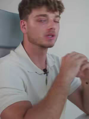 A medium shot features a fair-skinned man with curly brown hair and a light beard. He's wearing a white-colored polo shirt and is looking slightly off-camera with a focused expression, possibly speaking. A small microphone is visible clipped near his neckline. The background is plain and white, suggesting an indoor setting.