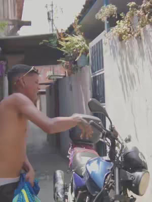 A young man, shirtless, stands next to a motorcycle, gesturing towards it. He is in an alleyway. The background includes a wall with plants and a partially visible building.