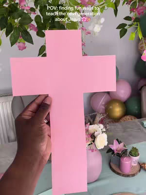 A hand holds up a large, pink paper cross in front of a background with green and light pink plant and balloon decorations, sitting on a blue tablecloth. The cross is the central focus, with the person's hand clearly visible.