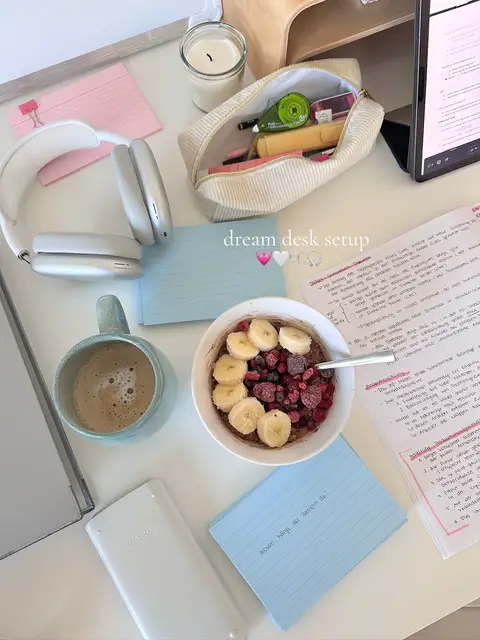 A top-down shot of a desk setup, a bowl of food with pieces of fruit, a coffee cup, headphones, and some study materials. Written: 'dream desk setup' with a pink and white heart.