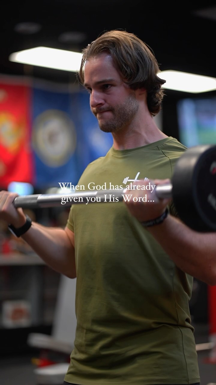 A man is in a gym holding a barbell, mid-lift, with text overlayed. He's wearing an olive green t-shirt and appears focused. The background is blurred, showing gym equipment and a flag.