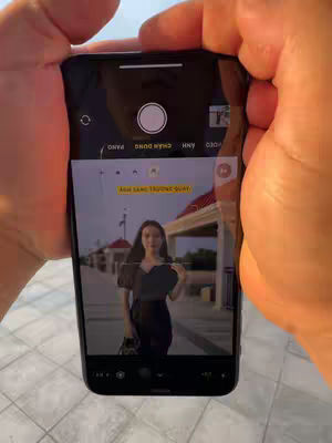An iPhone displays a portrait shot of a woman outdoors, held by hands. The image within the phone shows the woman in a dark dress posing on a walkway with buildings and sky in the background. A notification panel with yellow highlights is overlaid.