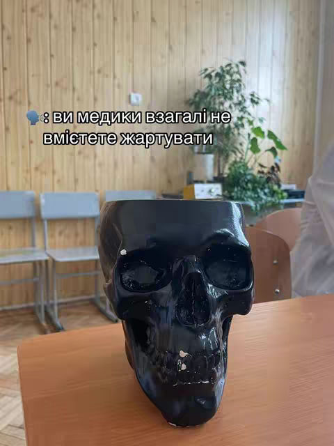 A black skull-shaped object is prominently displayed on a wooden table. The skull is highly polished, reflecting light in various areas. The background features a wooden wall and a green plant, suggesting an indoor setting. Text overlayed on the image features a cartoon thought bubble with text written in Ukrainian.