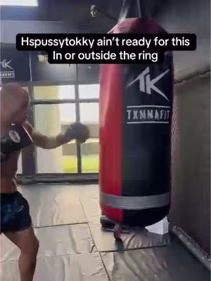 A man in a boxing gym practices punches on a heavy bag. The background is a blurred space with a window, while the foreground shows the man in boxing attire swinging his right fist at the bag.