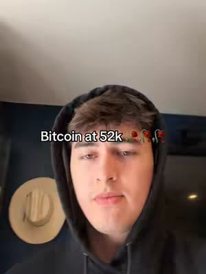 A young man wearing a black hoodie is centered in frame. The background features a white wall and part of a dark blue wall with a light-colored hat. Text overlay reads 'Bitcoin at 52k.' Colorful cartoon-style embellishments are above the text.