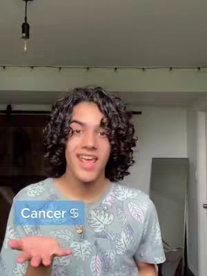 A young man with curly dark hair is shown from the chest up, his face centered with an open, friendly expression. He's wearing a light gray t-shirt with a subtle, repeating pattern. Behind him, a window and ceiling with a hanging light fixture can be seen. There's a semi-transparent blue box in the image with the word 'Cancer' and its astrological symbol.