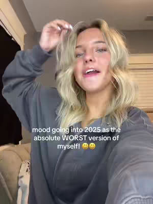A selfie-style video shows a young woman with blonde hair, slightly off-center, smiling and gesturing. The background is a blurry, neutral-toned room. Overlay text reads: 'mood going into 2025 as the absolute WORST version of myself!'