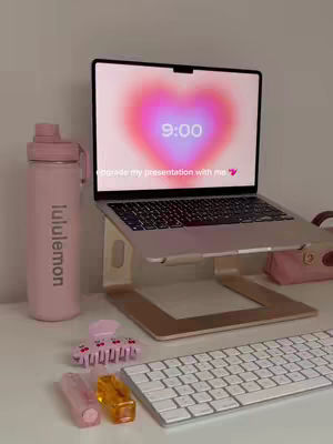 A well-lit workspace featuring a laptop open on a stand that displays an aesthetic heart-shaped background, along with various desk items like a pink water bottle, lipsticks, and a keyboard. The composition is clean and visually appealing, with a focus on soft, pastel colors.
