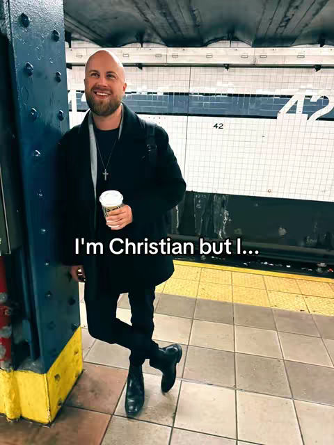 A man with a beard and a bald head stands in a subway station. He is wearing a black coat, black pants, and black boots. He holds a coffee cup in his hand. The background is a subway platform with a tiled wall.