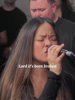 A woman with long brown hair is singing into a microphone with her eyes closed in front of a blurred background of people. Text overlayed reads "Lord it's been broken".