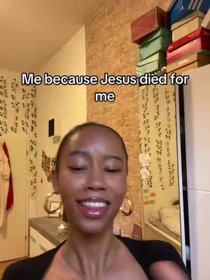 A woman with a smiling expression is centered in the frame. She has visible earrings and is wearing a dark top, with text overlayed that reads, "Me because Jesus died for me". The setting appears to be within a room with a door and stacked boxes visible in the background.