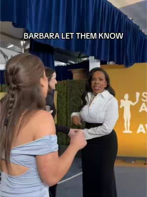 The image captures a moment at an award show. Two women are the focus. One is turned towards the other, who is center frame facing the camera and holding a microphone. The background features a golden wall with a logo and blue draping.