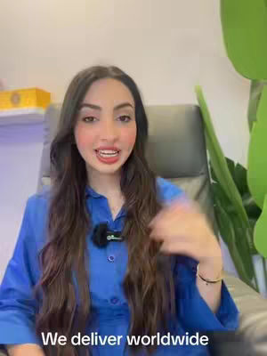 A woman with long, dark hair is seated in an office chair. She is wearing a blue button-down shirt. The text overlay reads 'We deliver worldwide'. The background includes a cream-colored wall, a green plant, and the edge of a shelf. She wears a small lavalier microphone.