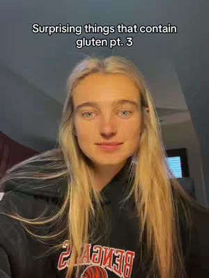 A medium shot captures a young woman with long blonde hair centered in frame, against a dark-toned background. Text overlay at the top reads, 'Surprising things that contain gluten pt. 3'. She wears a black hoodie. The composition focuses on her face and upper body, suggesting a direct address to the viewer with a serious but neutral expression.