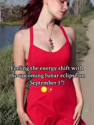 A woman in a red dress stands in front of a blurred background of a pond and trees. Text overlay on the image reads 'Feeling the energy shift with the upcoming lunar eclipse on September 17!' Below the text is a yellow circle and a star.