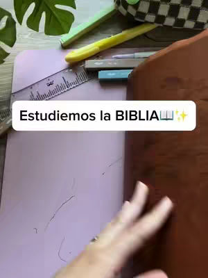 The image shows a person's hand holding a brown notebook with open pink pages. A ruler, pens, and a pencil case rest on top. The words "Estudiemos la BIBLIA" are written in a white box with sparkles. The background consists of a wooden table and some green leaves.