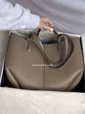 A hand holding a tan leather tote bag partially inside a white box with the text: the most perfect uni bag.