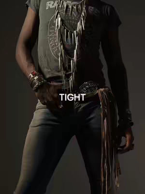 The image features a person from the waist up in a dimly lit setting. The person is wearing a grey t-shirt with a graphic, blue jeans, and a belt with a large buckle. The person has multiple bracelets on the left wrist and a detailed necklace. Text overlayed on the image reads 'TIGHT'.