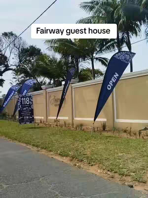 The video shows a guest house with the name "Fairway guest house" on a white sign. There are also flags saying open, breakfast, and displaying the number "118". The background shows green grass, trees, and sky. The road is visible in the foreground.