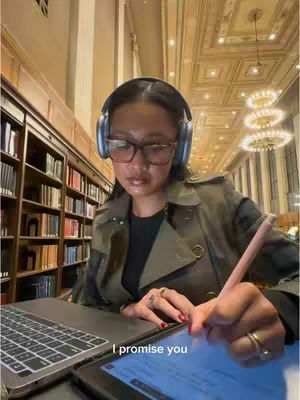 A woman wearing glasses and headphones sits in a library, writing on a tablet while a laptop sits in front of her. The library setting includes bookshelves and ornate ceiling lights.