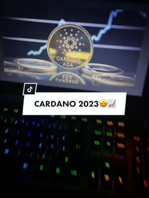 The image displays a physical Cardano (ADA) coin prominently in the center, slightly off-center. Behind the coin is a laptop screen showing a graph with a blue trend line heading upwards. Some other Cardano coins are also visible. In the foreground is a backlit mechanical keyboard with colorful keys. An overlay at the bottom indicates 'Cardano 2023'.