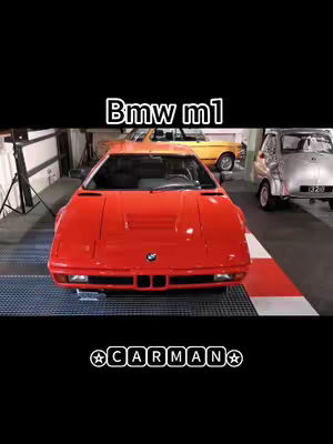 A red BMW M1 car is the subject of the image, positioned centrally with the front directed towards the viewer. The car is in a garage setting. The foreground is a black area. Below the car is text. Behind it a yellow car is parked