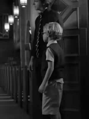 A black and white image shows two people in what appears to be a church. On the left, in the midground, several lights hang down. Standing in the foreground is a child wearing a vest, shirt and shorts, and glasses, facing the wall. To the child's right, an adult, in a suit, stands next to the child. The adult is at a slight angle.
