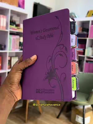 A hand holding a purple Women's Devotional & Study Bible, with colorful tabs. The background shows a partial bookstore interior.
