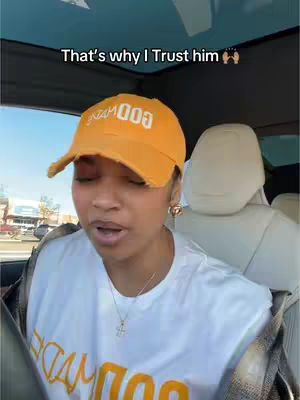 A woman wearing an orange cap and a white t-shirt is sitting inside a car. The background shows a car interior. The woman has a slight smile, and the composition focuses on her face and upper body.