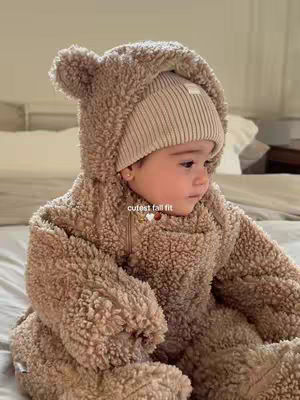 A baby wearing a light brown teddy bear outfit and a cream-colored beanie. The baby is sitting on a bed and looking to the side. The image is warm and inviting, with neutral tones.