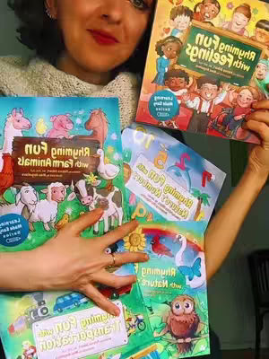 A woman with fair skin and brown hair is holding up a stack of colorful children's books, partially obscuring her face. The books feature bright illustrations and text, with titles like "Fun with Animals" visible.Her hands show a ring and her face shows a smile.