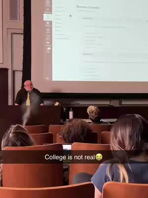 A medium shot captures a college lecture hall scene. The focus is on a professor standing behind a lectern. He is holding a cigarette that is emitting smoke, as if it's been lit mid-lecture. The foreground includes the backs of students' heads in the audience, with rows of chairs and what seems to be a projector screen in the background. The text overlay says 'College is not real'.