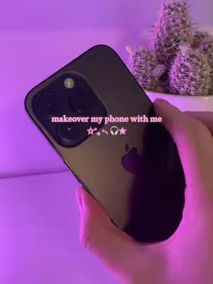 An iPhone is held at an angle with the camera lenses facing toward the viewer. A hand holds the phone, and the background is blurred with a purple hue. The text overlay reads, "makeover my phone with me" with multiple symbol emojis.