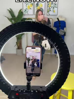 A woman is setting up for a live video, framed by a ring light. She's holding up what appears to be a phone while standing in front of a boutique setting. The scene is well-lit, likely to enhance the image.