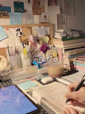 A well-lit study desk, featuring a laptop displaying a bright screen, handwritten notes, pens, and a mug of a beverage. The background includes a corkboard with notes and decorative items, alongside books and other stationary.