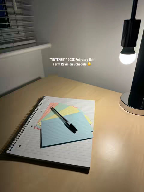 A dimly lit desk with a notepad, colorful sticky notes, and a pen. A black reading lamp casts a pool of light on the scene from the right.